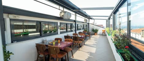 Un área de comedor al aire libre con varias mesas y sillas, amplias ventanas y plantas en macetas.