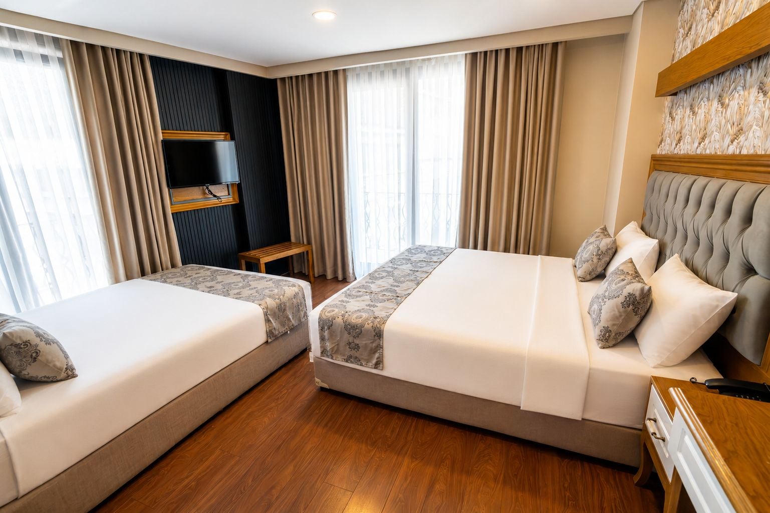 A hotel room with two beds, a wooden floor, a large window with curtains, and a wall-mounted shelf.