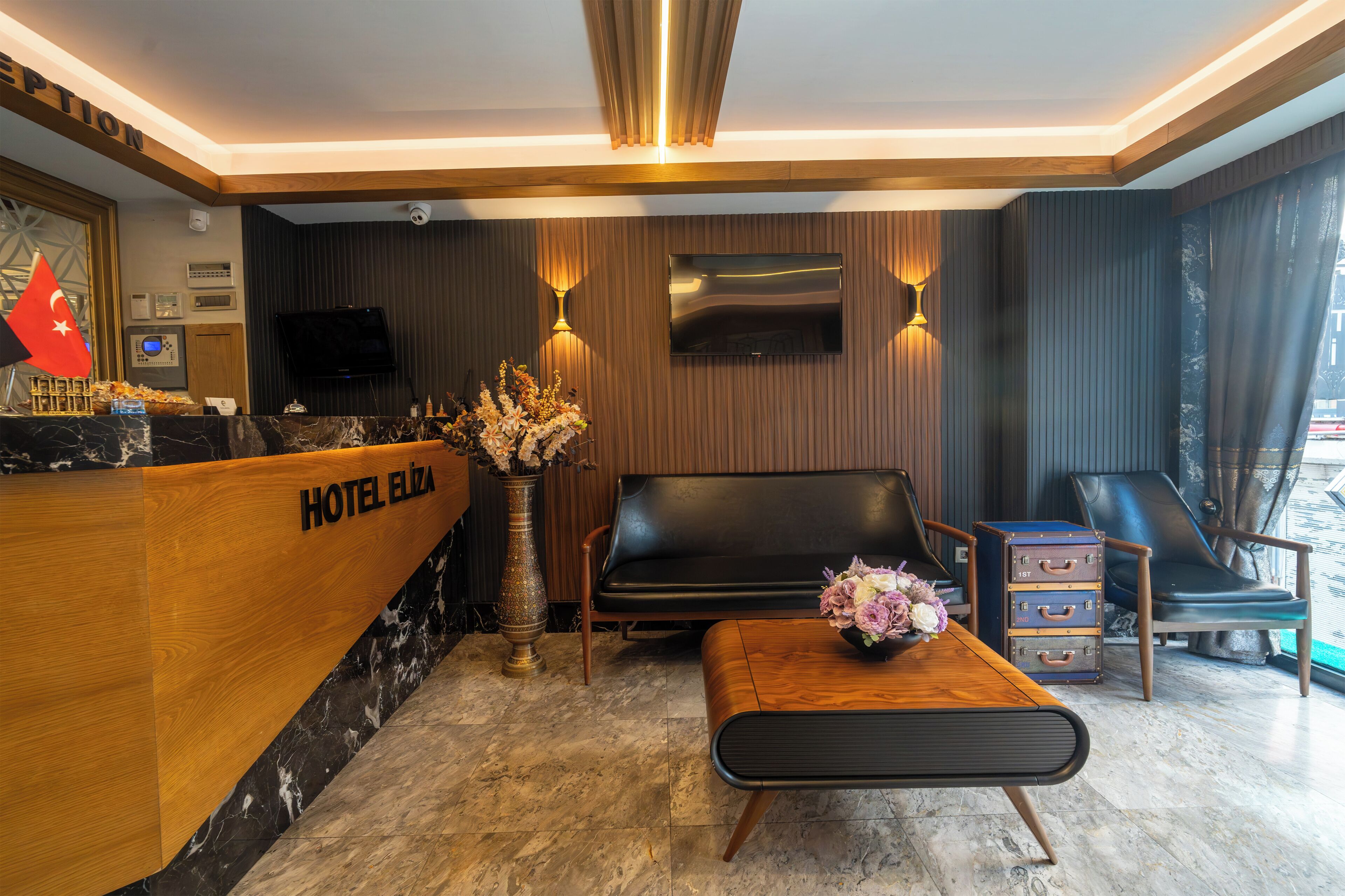 Een hotel receptie met een houten receptiebalie, een zwarte leren bank, een klein houten tafeltje met een bloevas, en een zithoek met twee stoelen.