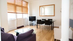 A modern living room with a glass coffee table, a flat-screen TV, a dining area with a table and chairs, and large windows with blinds. - Modern 2 bed flat in historic building (London)