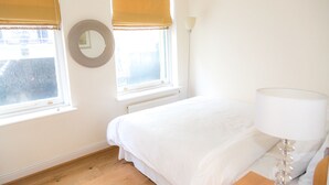 A bedroom with a bed, a round mirror, two windows with blinds, a radiator, and a bedside lamp. - Modern 2 bed flat in historic building (London)
