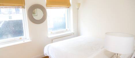 A bedroom with a bed, a round mirror, two windows with blinds, a radiator, and a bedside lamp.
