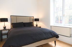 A bedroom with a large bed, two bedside tables with lamps, a window with a view of buildings, and a radiator under the window. - Modern 2 bed flat in historic building (London)