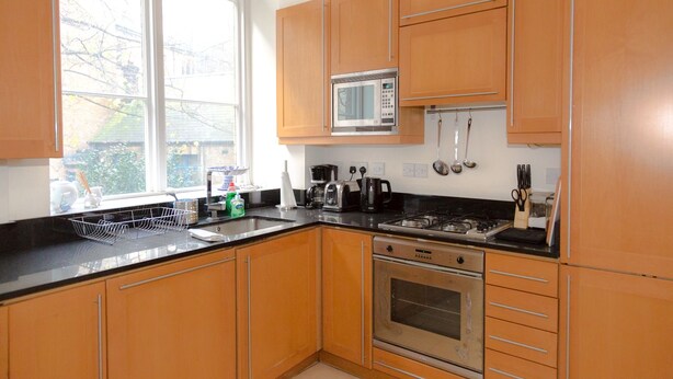 A kitchen with wooden cabinets, a microwave, a stove, and a window with blinds. - Modern 2 bed flat in historic building (London)