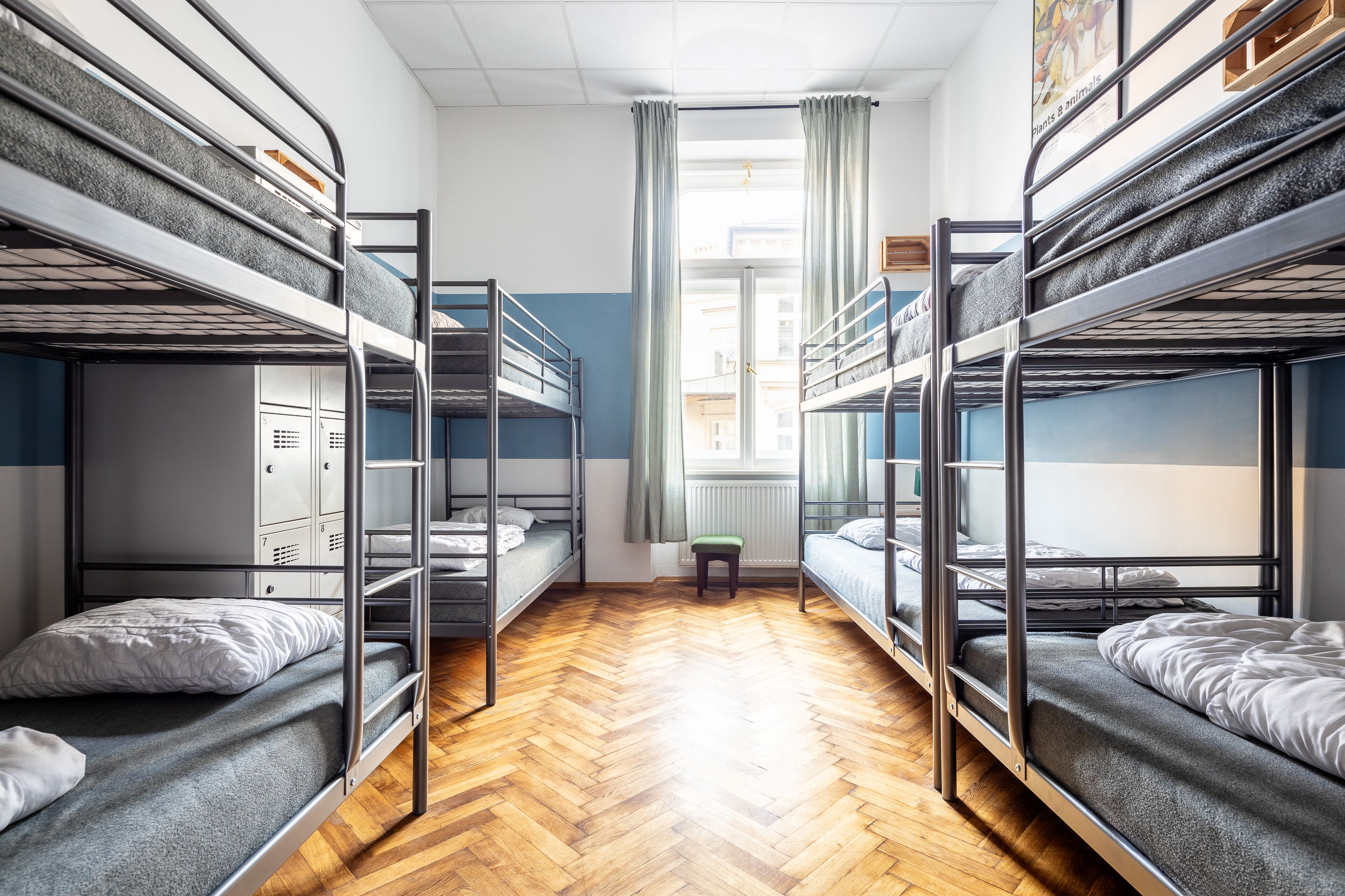 Une chambre de dortoir avec des lits superposés, un parquet en chevrons, un radiateur et une fenêtre avec des rideaux.