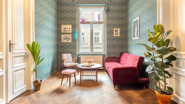 A room with a herringbone wooden floor, a red sofa, a small round table with a vase, and a potted plant. - Das Hostel (Kraków)