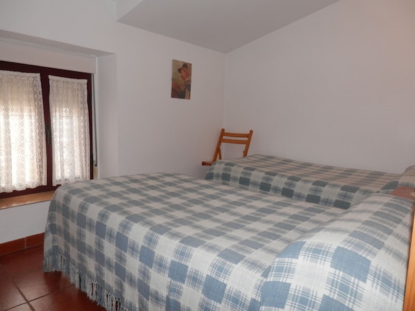A small, simple room with a bed, a wooden chair, and a window with curtains.