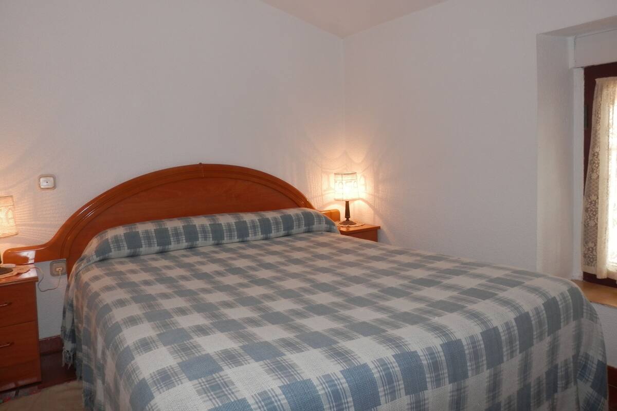 A bedroom with a wooden headboard, a checkered bedspread, two bedside lamps, and a nightstand with a lamp.