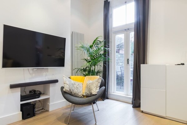 A modern living room with a flat-screen TV, a grey armchair with patterned cushions, a potted plant, and a sliding glass door leading to an outdoor area.