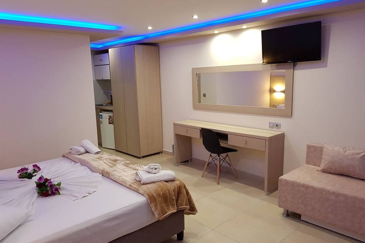 A hotel room with a bed, a desk, a chair, a TV, and a kitchenette.