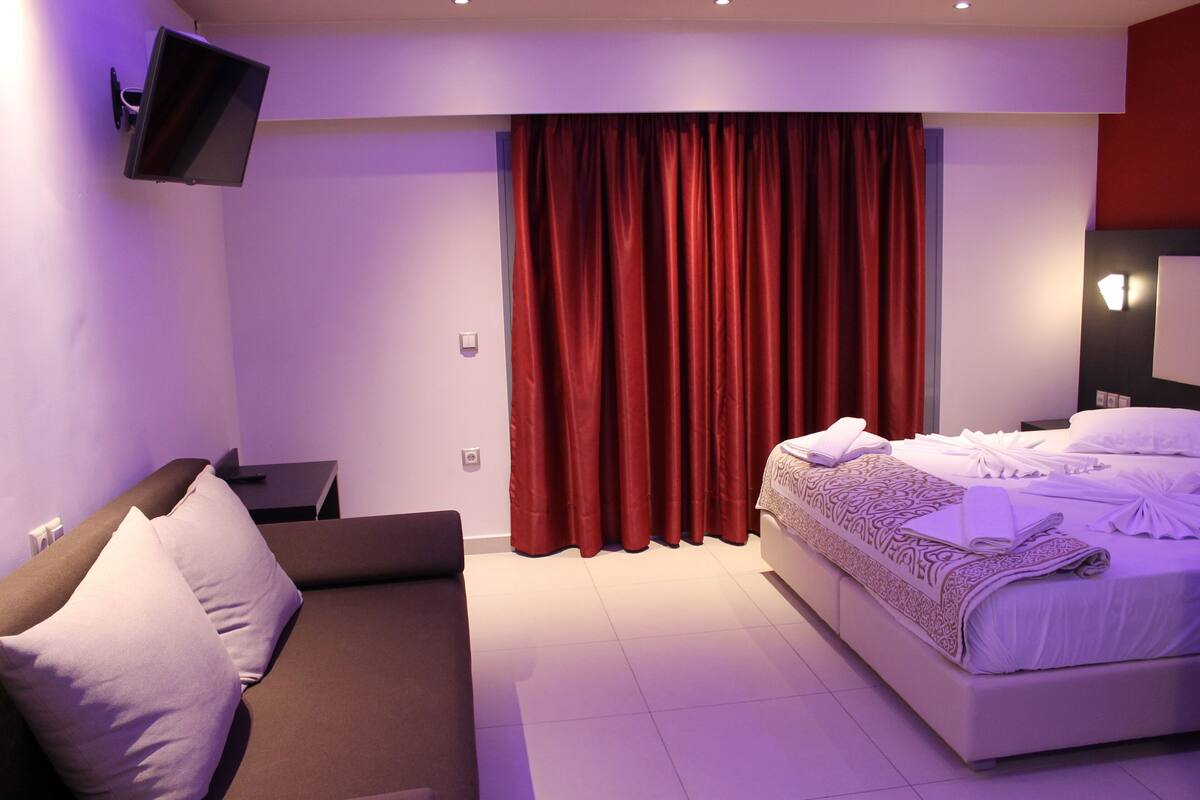 A modern hotel room with a bed, a sofa, a flat-screen TV mounted on the wall, and red curtains.