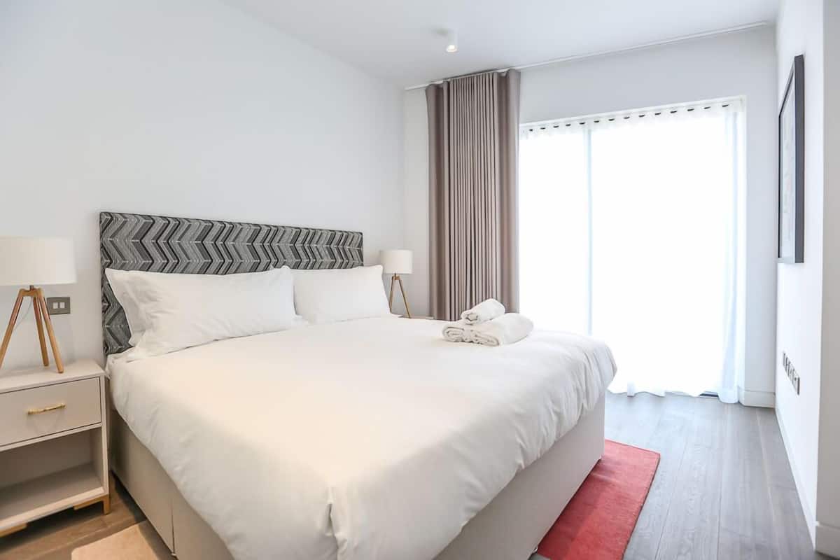 A modern hotel room with a large bed, a bedside table with a lamp, and a view of the outside through a window with curtains.