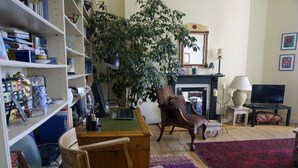 Ein Wohnzimmer mit Bücherregal, Schreibtisch, einem Sessel mit Leopardenmuster, einer großen Pflanze, einem Kamin und einem Fernseher.