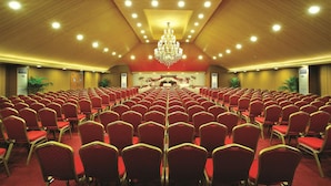A large conference room with rows of red chairs, a chandelier, and a stage with floral decorations. - The Classik Fort (Kanayannur)
