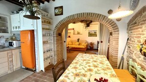 A kitchen with an orange refrigerator, a dining table with a floral tablecloth, and a brick archway leading to a living room. - Hous Malerin with sea view, fenced garden - by ToscanaTour (Guardistallo)