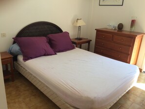 Une chambre à coucher avec un lit, des oreillers violets, une commode en bois, une table de chevet, une lampe et un vase posé sur la commode.