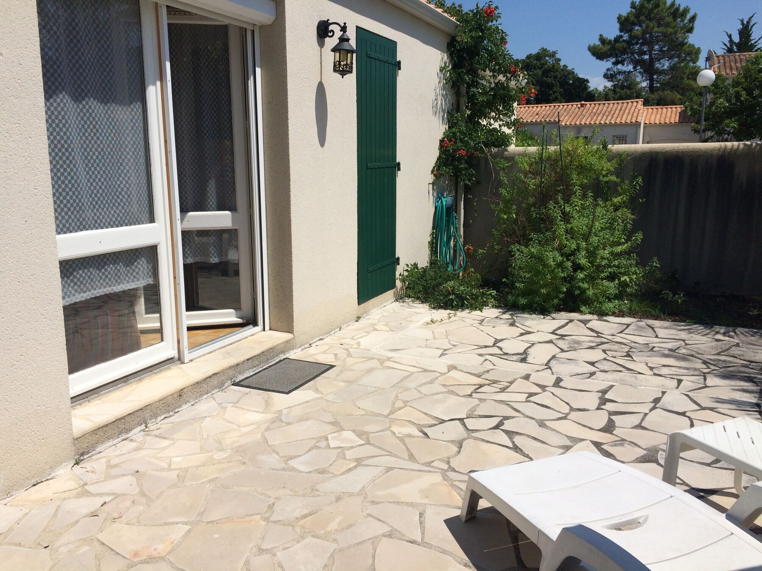 Une terrasse avec un sol en pierre, une porte-fenêtre coulissante, une porte verte et une petite table blanche.