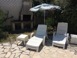 A patio with two white lounge chairs and a small table, an umbrella, and a stone-tiled floor.