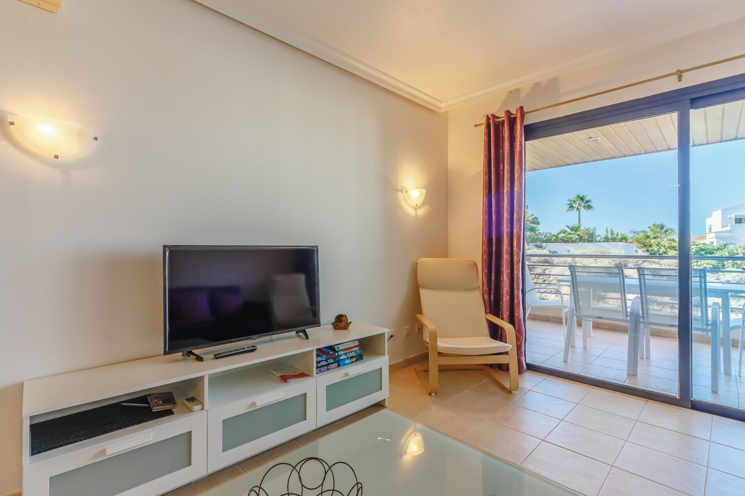 A modern living room with a flat-screen TV, a white entertainment unit, a glass coffee table, a beige armchair, and a sliding glass door leading to an outdoor patio with chairs.