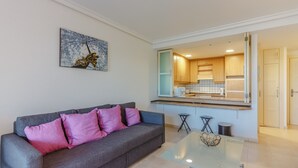 A modern living room with a grey sofa, pink cushions, and a glass coffee table. A kitchen is visible through an open door, featuring wooden cabinets and a tiled backsplash. - Balcon de Los Gigantes 2A037 (Acantilados de Los Gigantes)