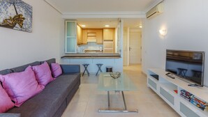 A modern living room with a grey sofa, a glass coffee table, a TV on a white stand, and a kitchen in the background. - Balcon de Los Gigantes 2A037 (Acantilados de Los Gigantes)
