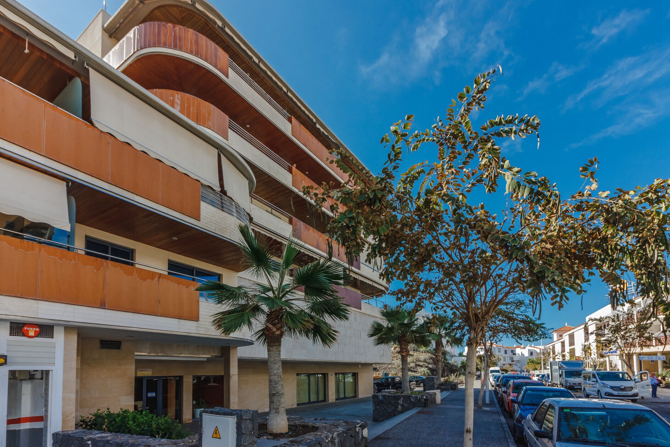 A multi-story hotel with balconies, palm trees, and parked cars.