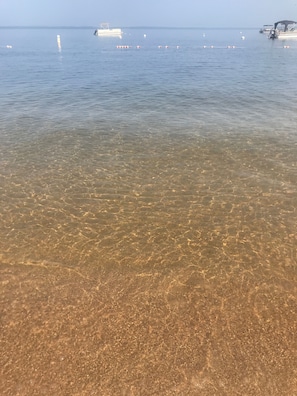Klar udsigt over en strand med roligt vand, en båd og bøjer i vandet.