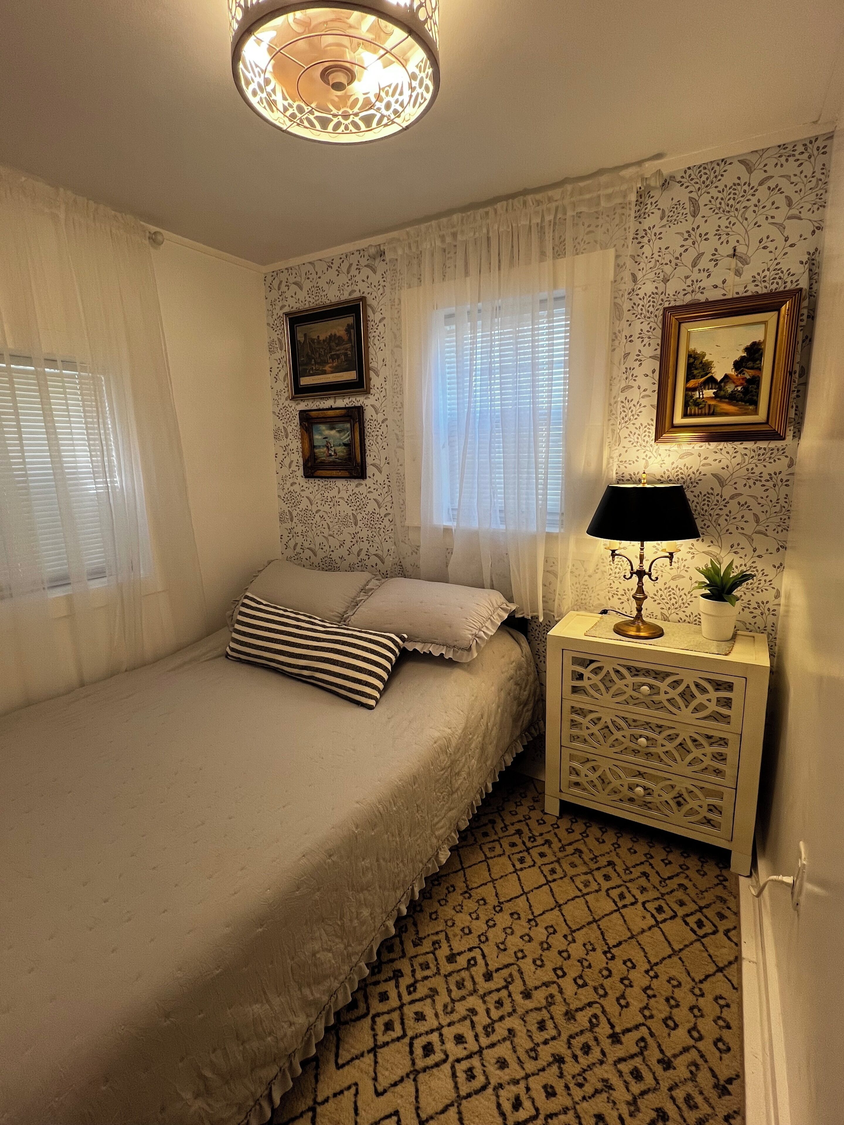 A bedroom with a bed, a nightstand with a lamp, two framed pictures, and patterned wallpaper.