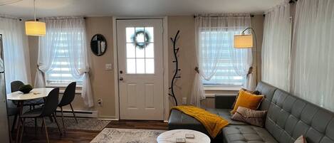 A living room with a sofa, a coffee table, a dining area with chairs, a chandelier, and a door with a wreath.