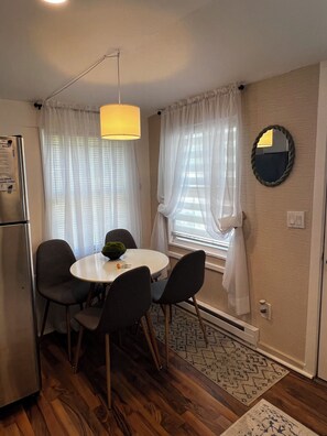 A compact kitchen with a refrigerator, a dining table with chairs, a hanging light fixture, and a window with curtains. - COZY,Newly renovated,West Shores ,short walk to Nasons Beach 🏖🏠 (Sebago)