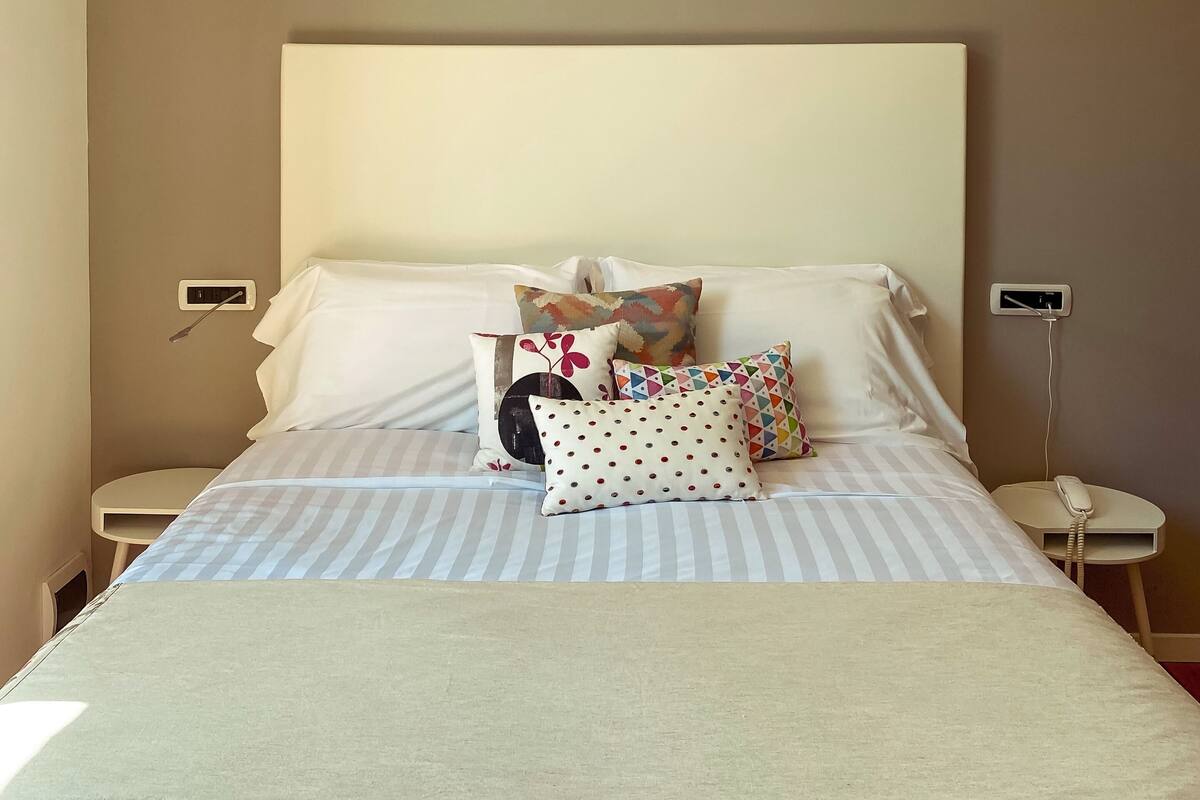 A neatly made bed with striped bedding and multiple pillows, including a polka-dotted one. There are bedside tables with a phone and a power outlet on each side.