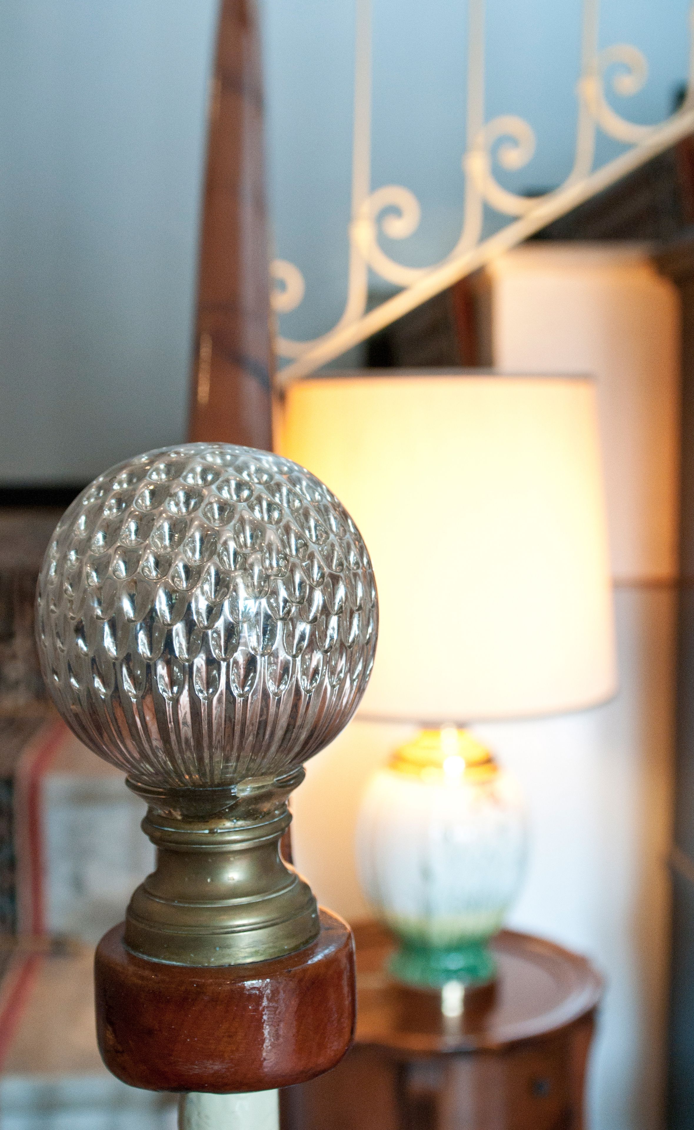 A close-up of a decorative glass knob on a wooden base, with a blurred background featuring a lamp and a patterned wall.