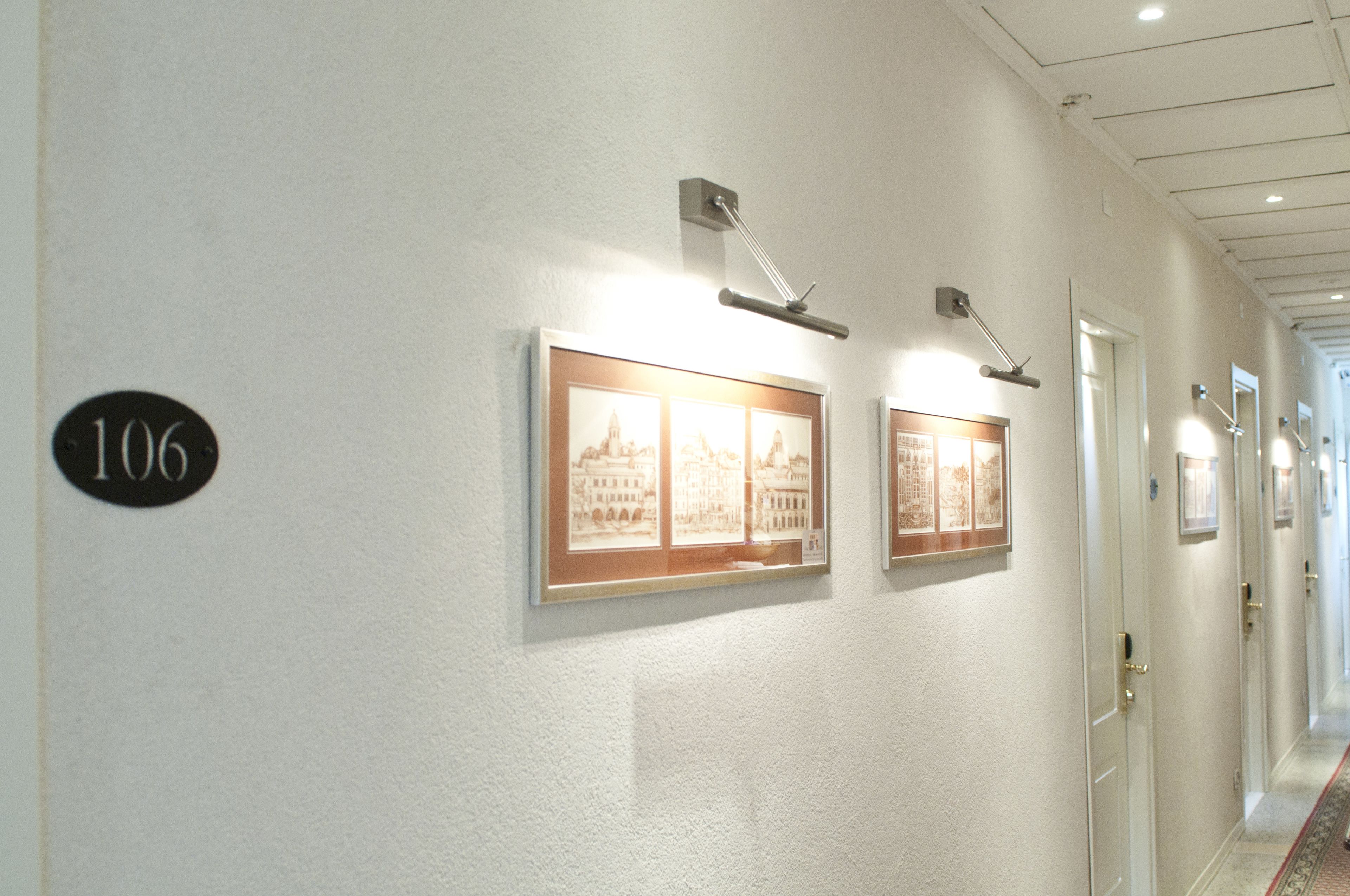 Un couloir avec des tableaux encadrés accrochés au mur, une porte portant l’inscription « 106 », et un sol recouvert de moquette.