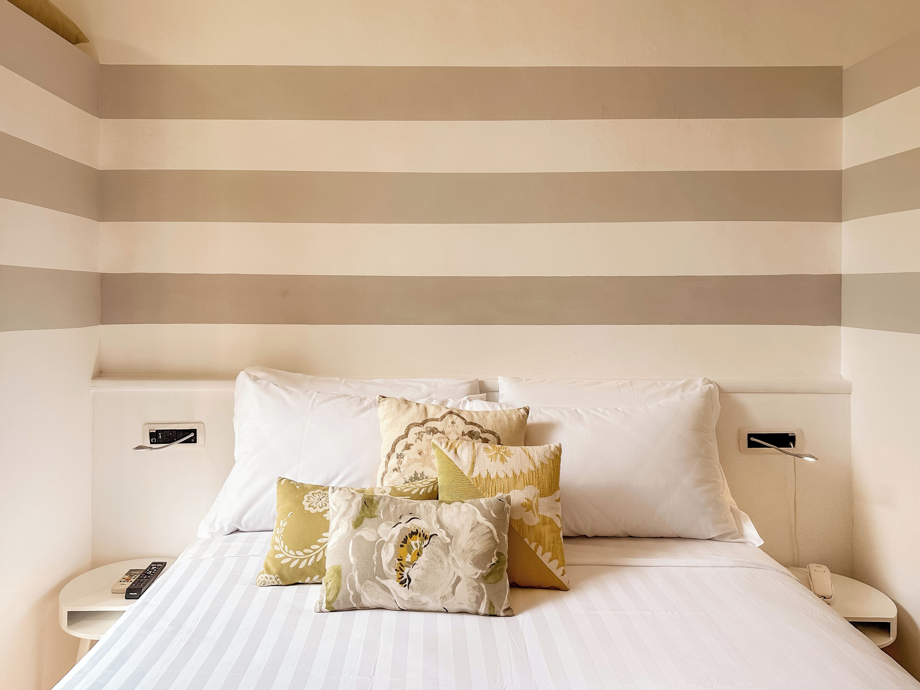A hotel room with a striped wall design, a neatly made bed with multiple pillows, and bedside tables with remote controls.