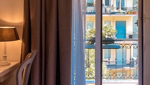 A room with a bed, a chair, a table, and a view of a balcony with potted plants and a blue shutter.