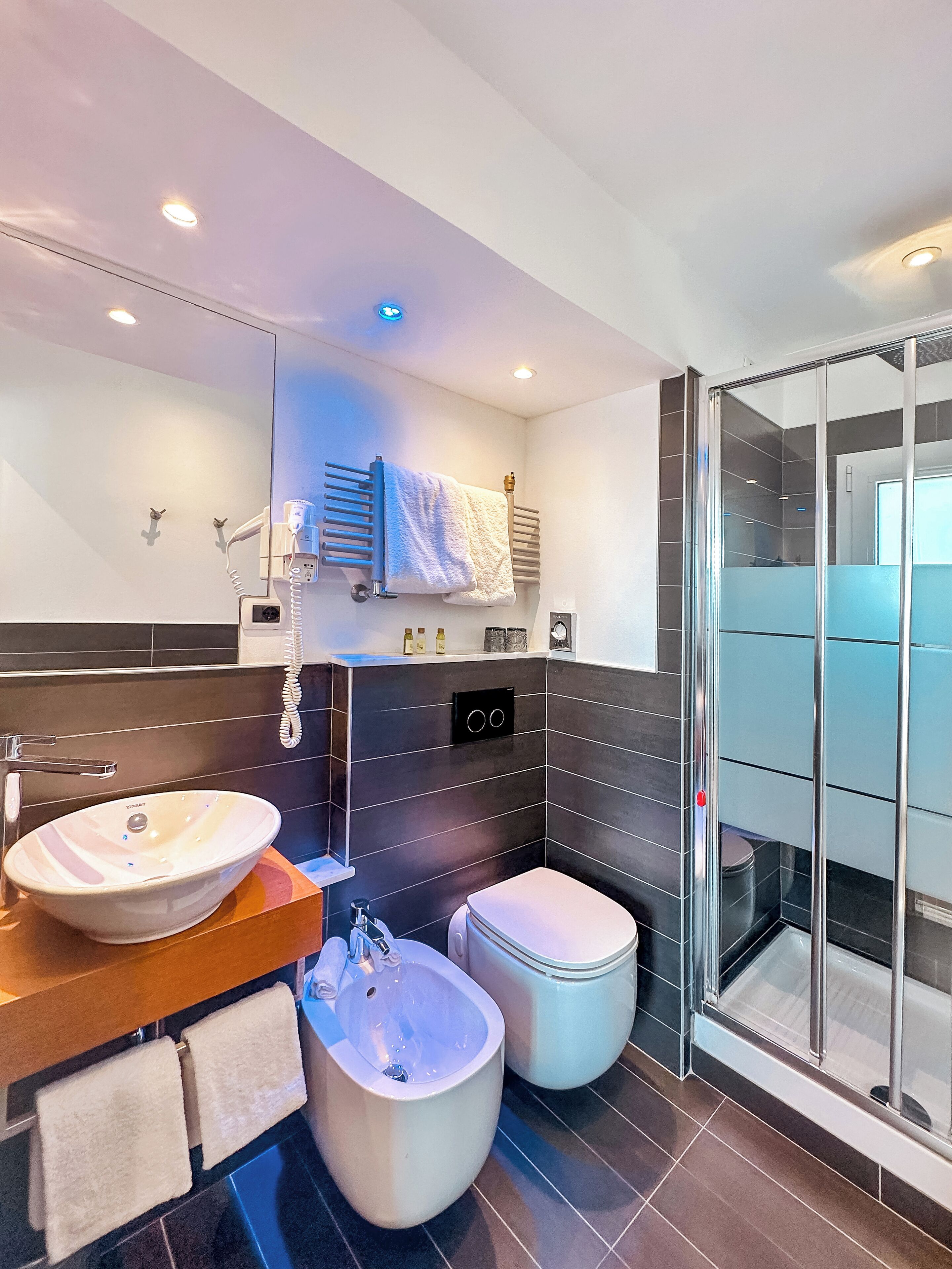 Ein modernes Badezimmer mit einem großen Waschbecken, einem Bidet und einer Toilette, einer Duschkabine aus Glas sowie einem Handtuchhalter mit Handtüchern.