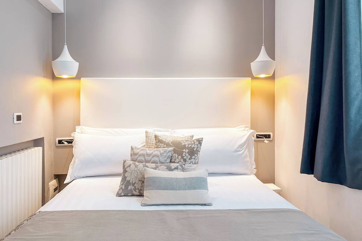 A neatly made bed with a white duvet, multiple pillows, and a bedside table with a lamp.