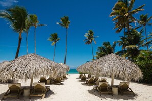 A beach with thatched umbrellas and lounge chairs, palm trees, and a clear blue sky.