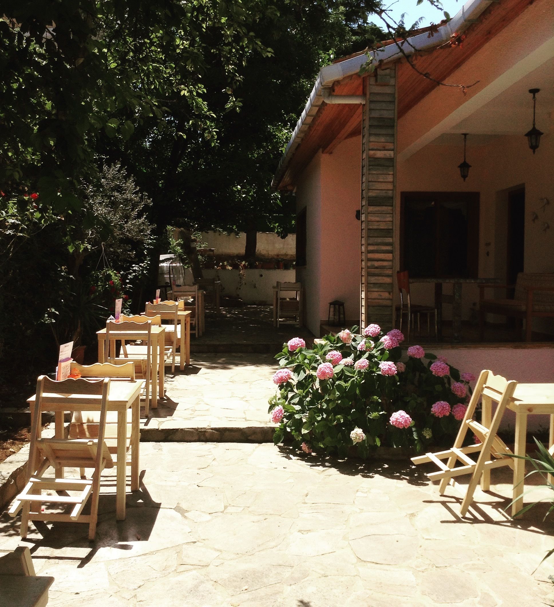 An outdoor patio with wooden tables and chairs, a stone floor, and a pink flowering plant.