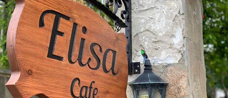 A wooden sign with "Elisa Cafe" written on it, mounted on a stone wall with a black lantern beside it.
