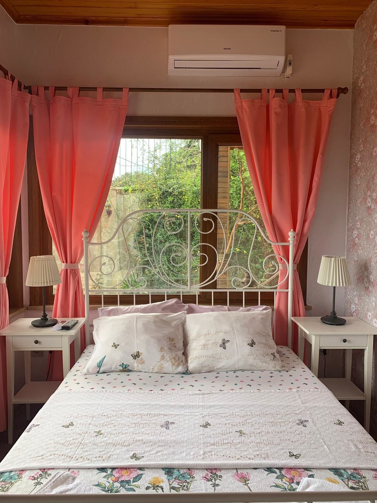Un dormitorio con cama, dos mesitas de noche, una ventana con cortinas, un equipo de aire acondicionado y vistas al exterior con vegetación.