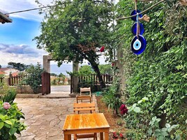 Un patio de piedra con mesa y sillas de madera, rodeado de exuberante vegetación y vistas al mar.