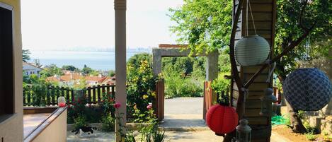 A covered outdoor area with a stone pathway leading to a wooden gate. There are potted plants and flowers, and a view of a cityscape and body of water in the distance.