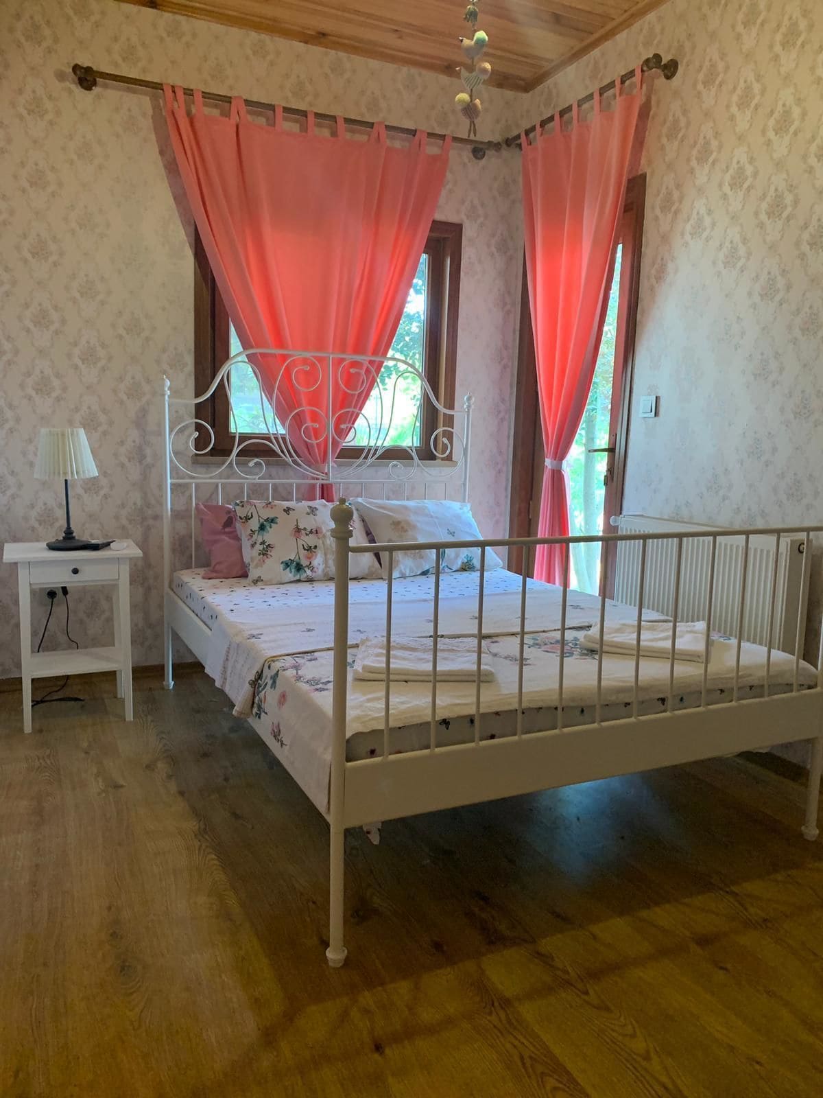 Ein Schlafzimmer mit weißem Metallbettgestell, blumigem Bettbezug, einem Nachttisch mit Lampe und rosa Vorhängen.