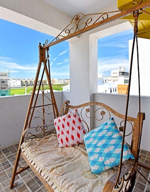 A balcony with a swing chair, two patterned cushions, and a view of buildings and greenery.