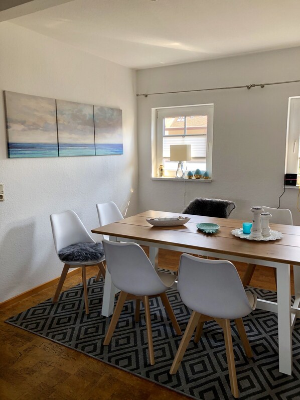 A dining area with a wooden table, white chairs, a patterned rug, a painting on the wall, and a window with blinds.
