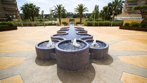 A series of circular water features arranged in a symmetrical pattern on a tiled patio, with palm trees and a building in the background. - MALDIVAS Apartments (La Pineda)