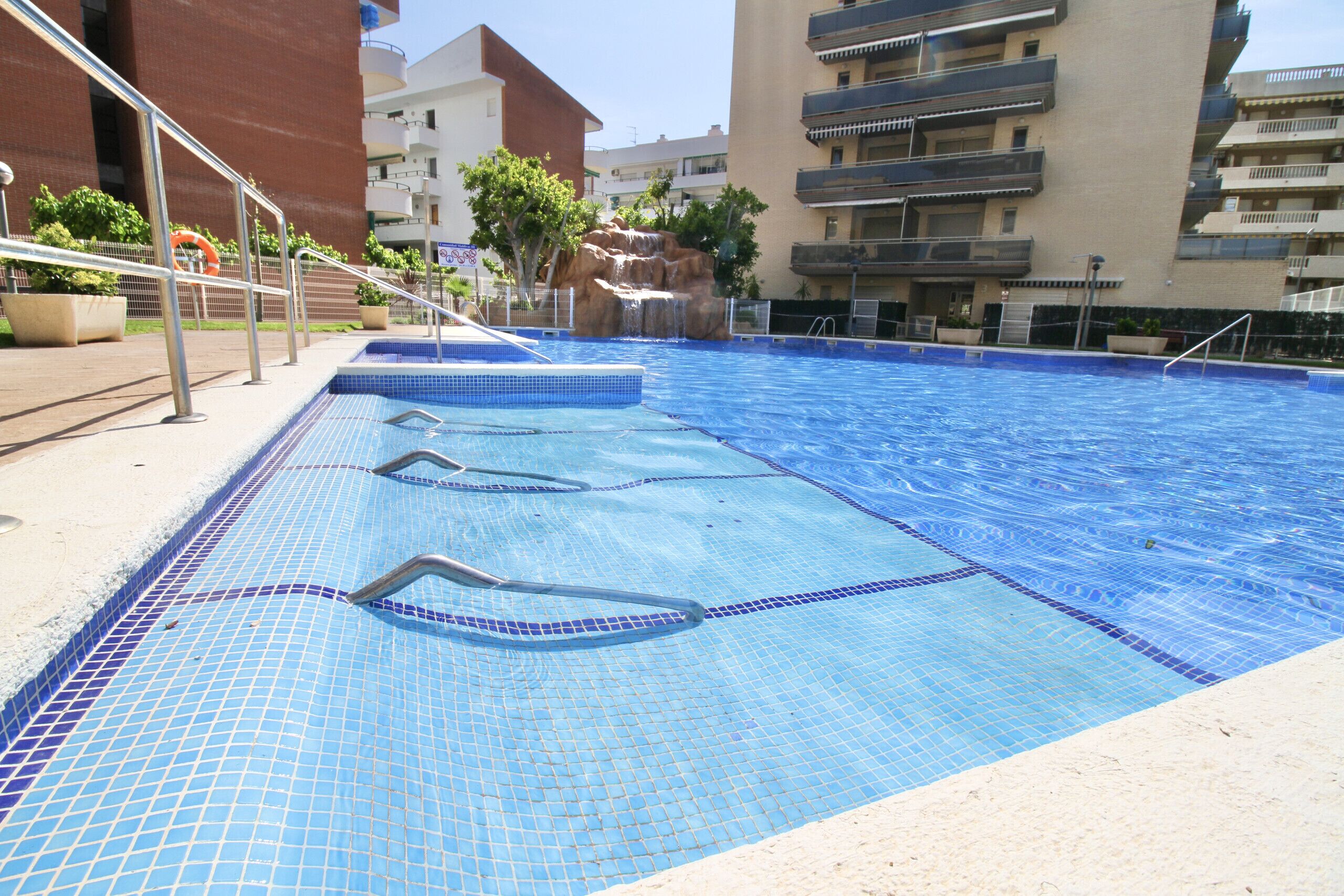 A swimming pool with clear blue water, surrounded by a sandy deck.