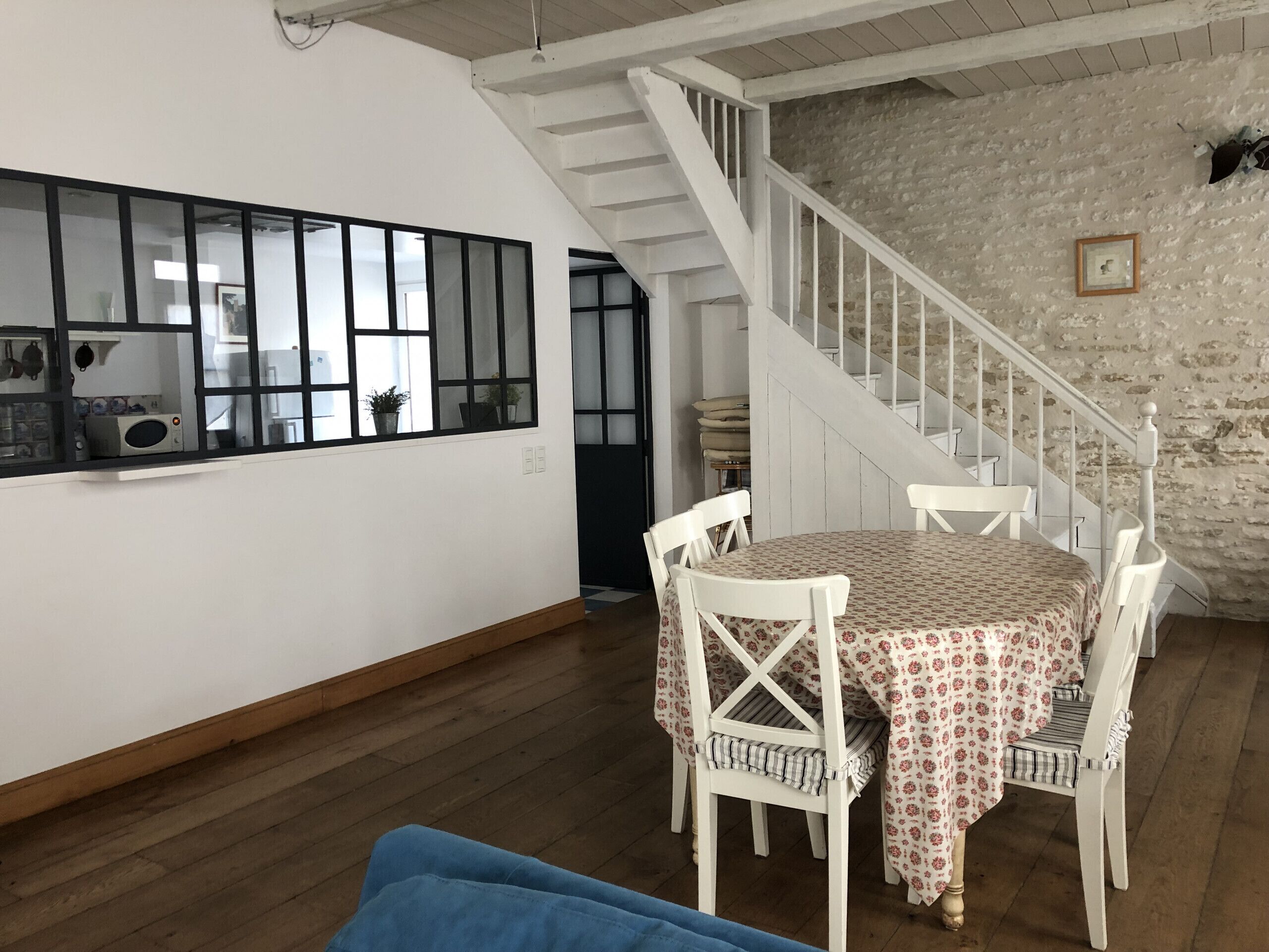 Un coin repas avec une table dressée pour quatre personnes, des chaises blanches, un plancher en bois, un escalier avec des rampes blanches et un mur en pierre.
