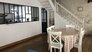 Un comedor con una mesa dispuesta para cuatro, sillas blancas, suelo de madera, una escalera con barandillas blancas y una pared de piedra.